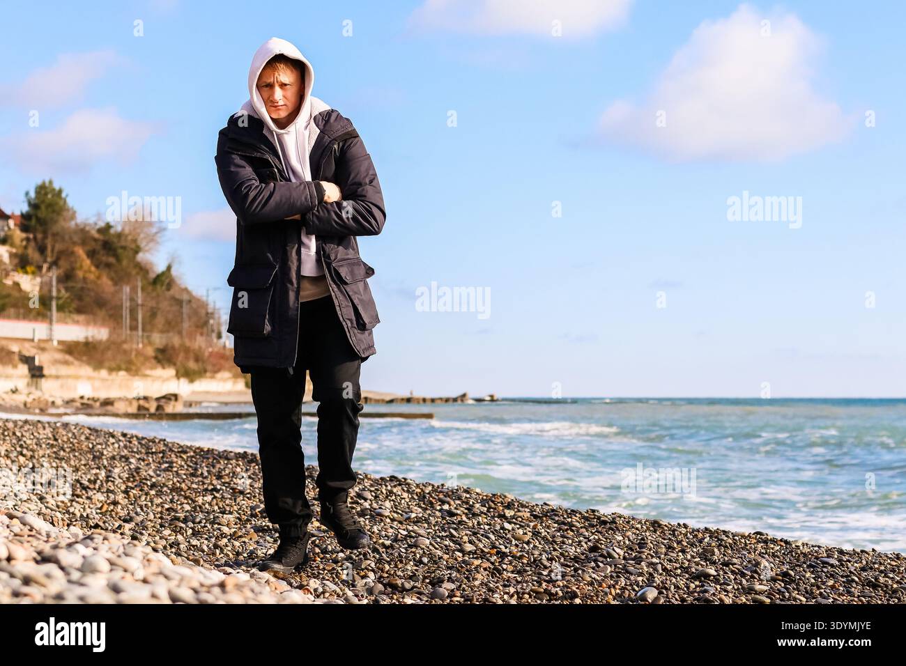 Erwachsener Mann in dunklem Mantel und heller Kapuze überkreuzt die Arme am steinigen Strand mit Meerblick unter teilweise bewölktem Himmel Stockfoto