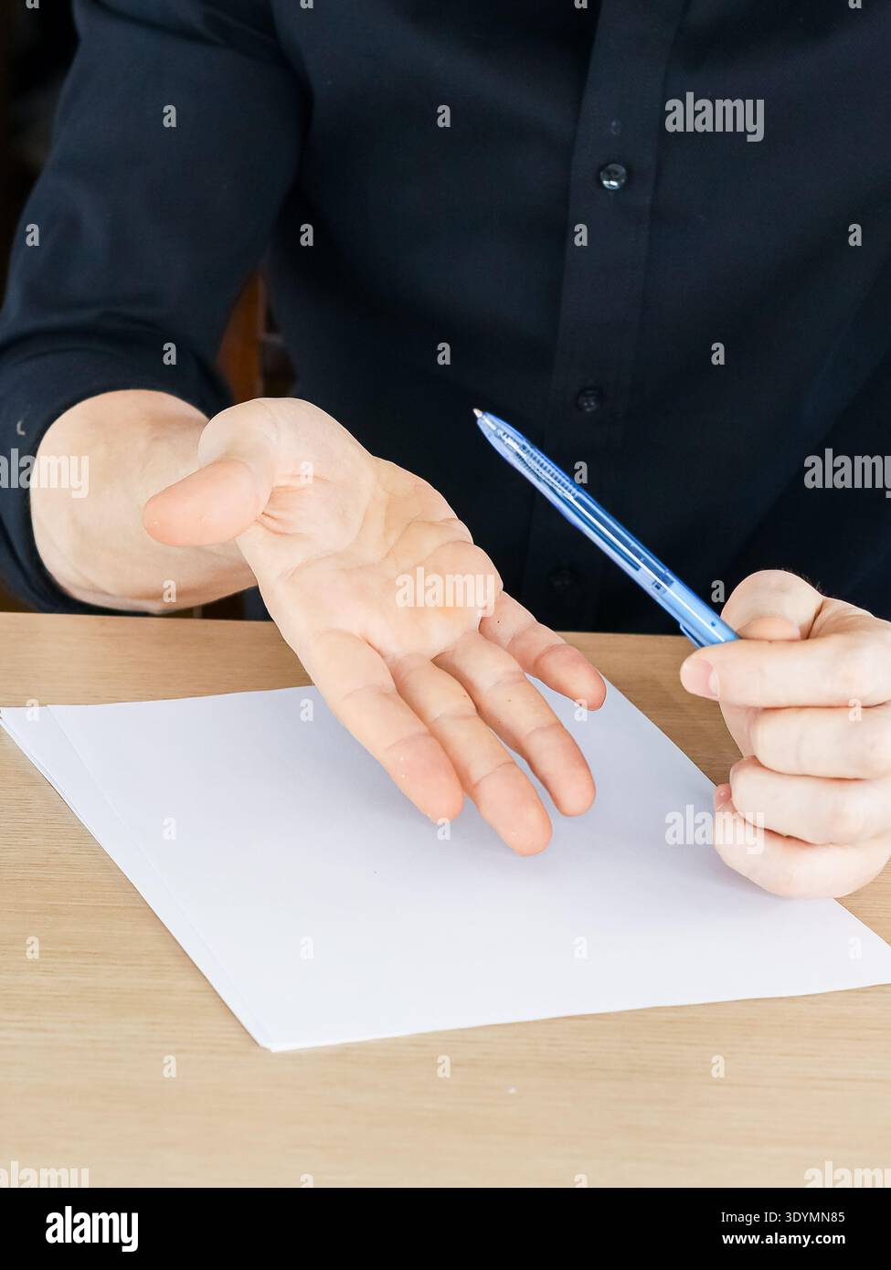 Männliche Hände über Holztisch, rechte Faust hält den blauen Stift nach oben, linke Handfläche zeigt nach oben in der Nähe eines weißen leeren Papierblatts Stockfoto