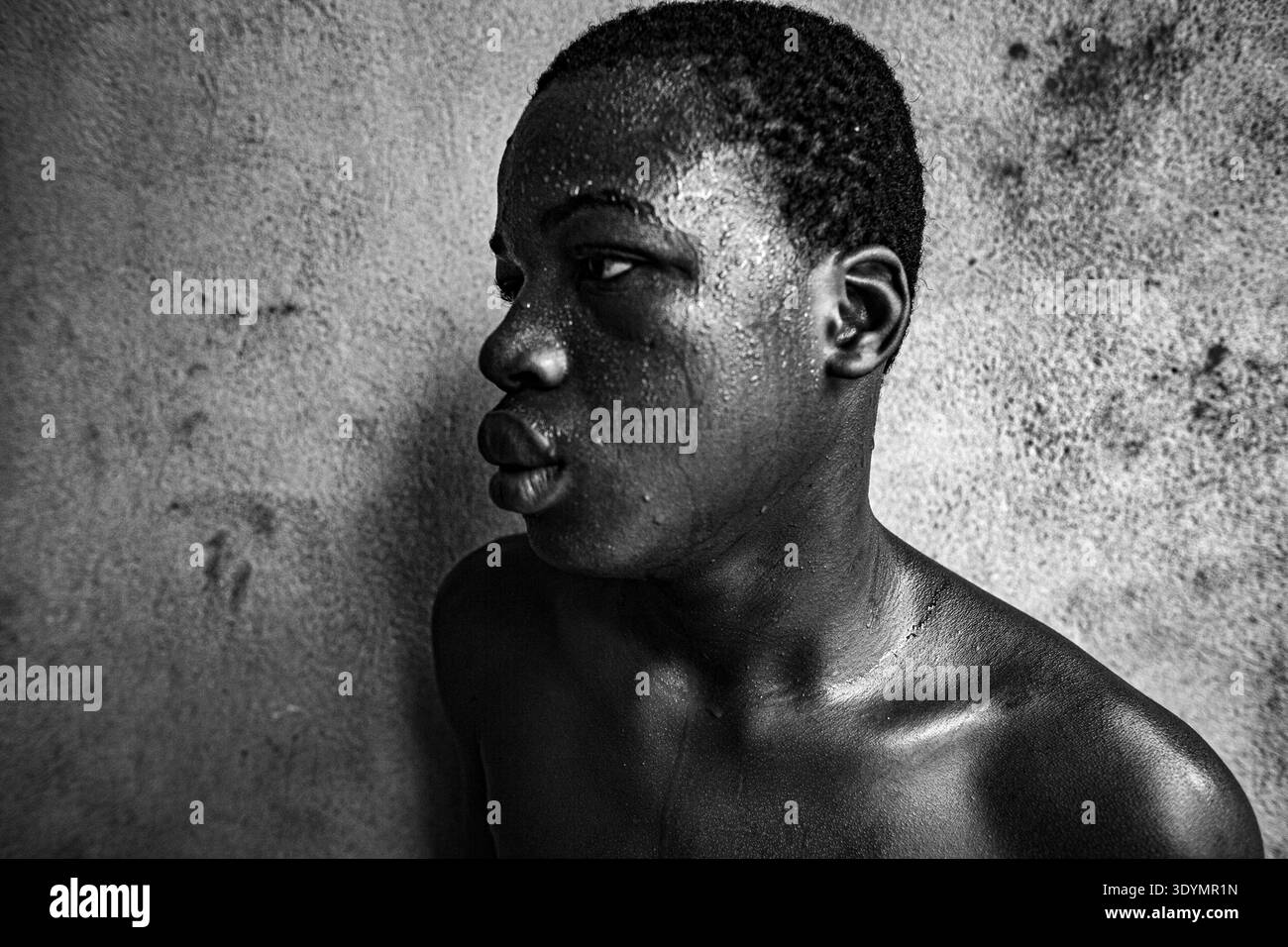 Schwarz-weiße Nahaufnahme-Porträt eines jungen afrikanischen Tänzers, der während einer erholsamen Pause mit Schweiß bedeckt ist und einen Moment ruhiger Intensität reflektiert. Stockfoto