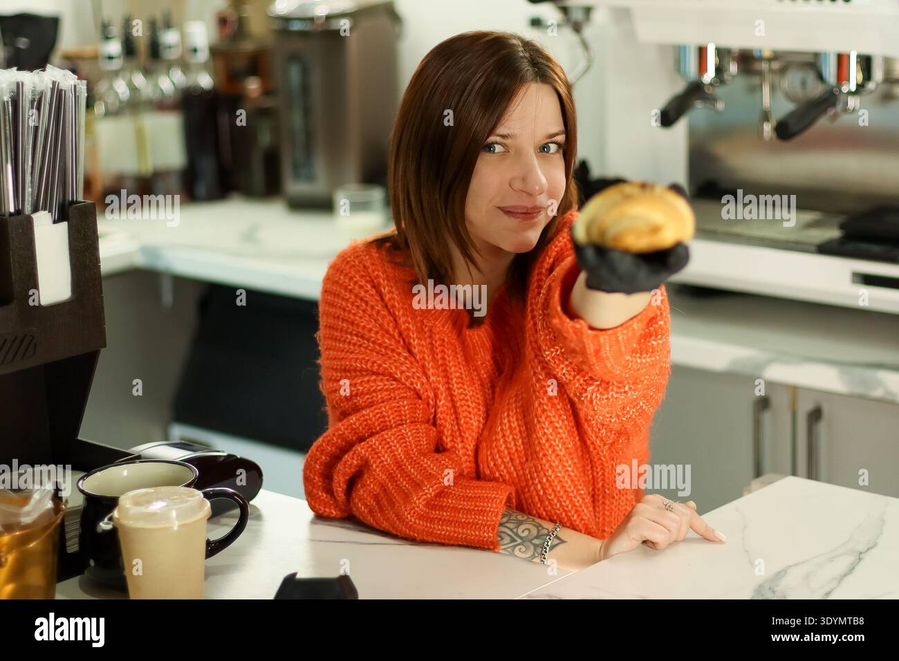 Der Barista in einem gemütlichen orangefarbenen Pullover hält frisch gebackenes Croissant neben Tassen und Ausrüstung auf weißem Marmor. Stockfoto