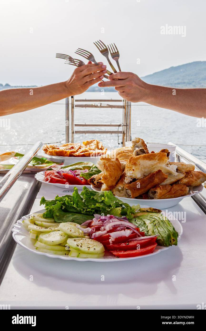 Eine Gruppe von Freunden genießt eine gemeinsame Mahlzeit auf einer Yacht und klickt in einer verspielten Geste über Tellern mit frischem Gemüse und Gebäck die Gabeln. Stockfoto