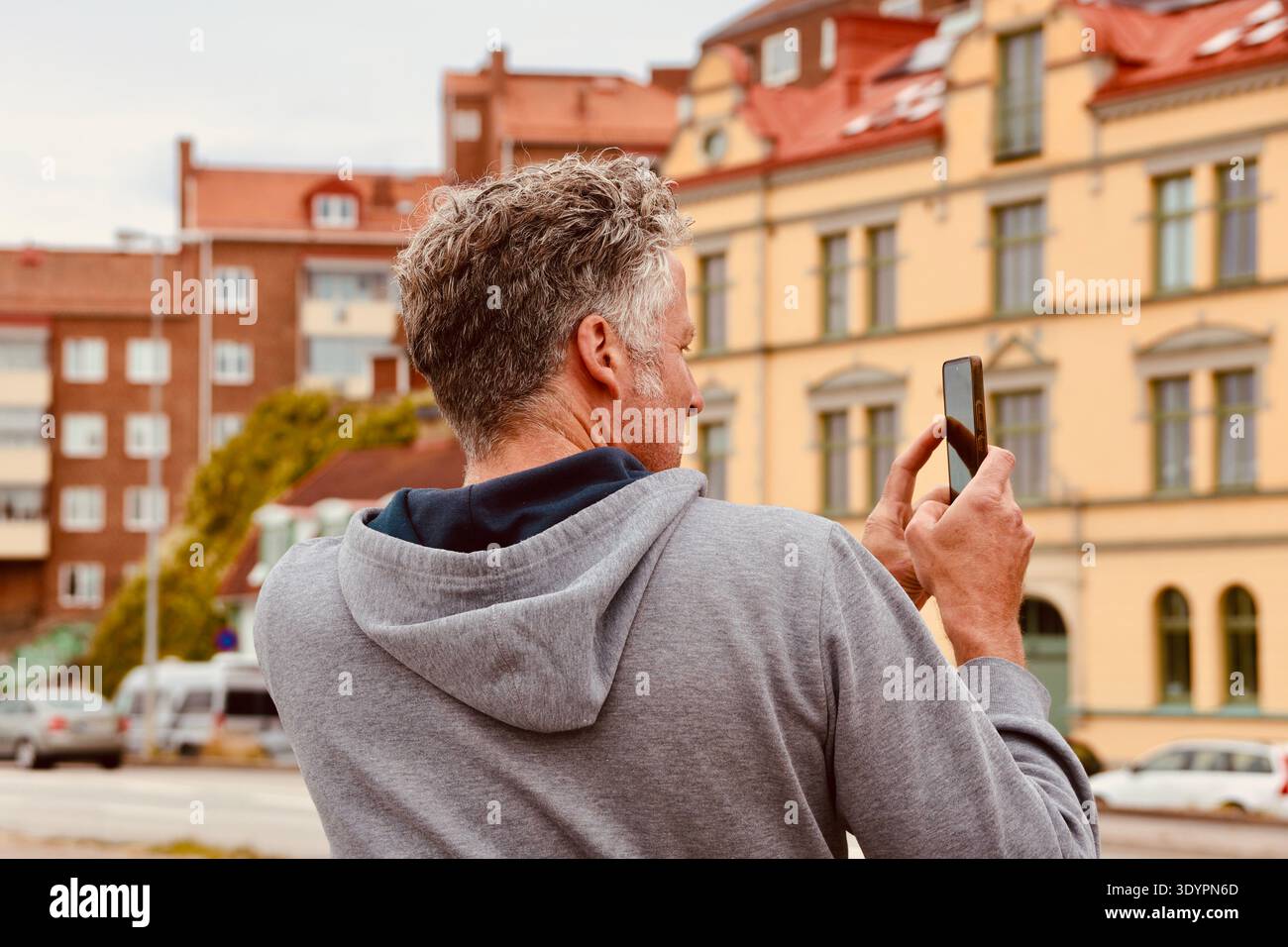Erwachsener Mann, der ein Smartphone benutzt, um ein Foto zu machen, während er in einer europäischen Stadtstraße mit farbenfrohen historischen Gebäuden im Hintergrund steht. Stockfoto