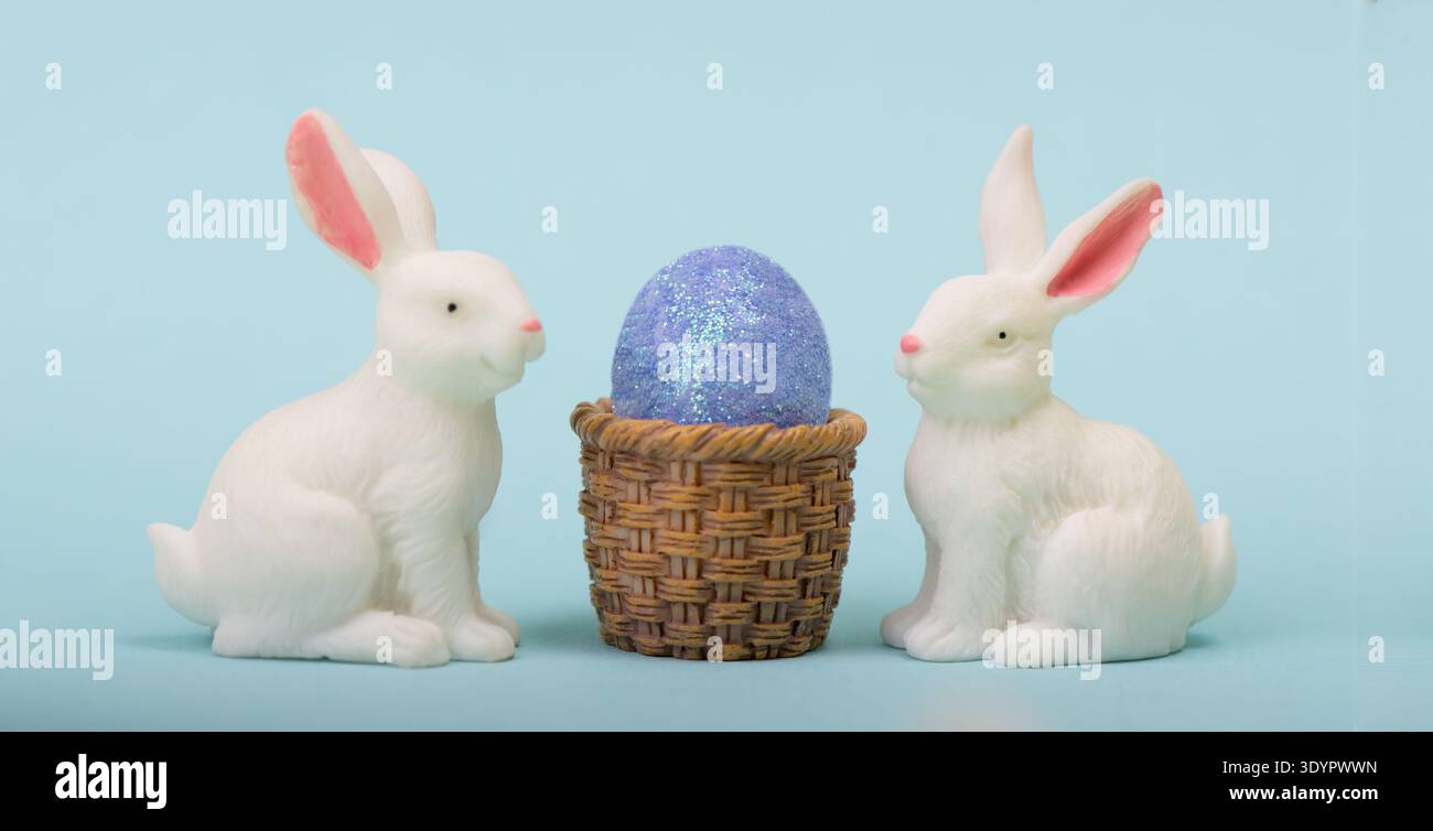 Zwei weiße Osterhasen-Figuren vor blauem Hintergrund mit Osterei und dem Korb-Kopierraum. Gut für Ostergrüße, Bannerdesign Stockfoto