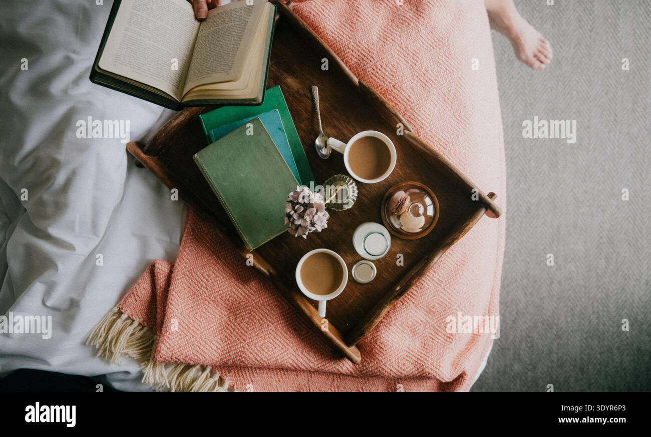 Auf einem Holztablett können Kaffee, Kerzen und Bücher auf einer gemütlichen rosa Decke aufbewahrt werden. Das offene Buch lädt zum Lesen oder zur Ruhe am Morgen ein. Das Tablett liegt neben dem weichen weißen Bett Stockfoto