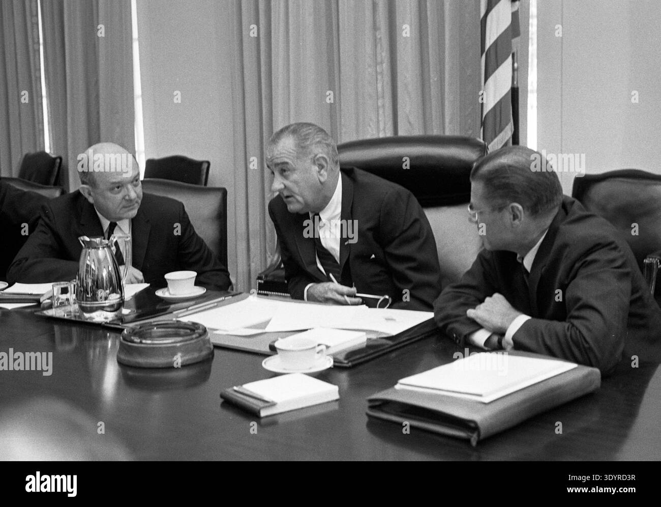Foto mit Verteidigungsminister Robert McNamara (rechts), Präsident Lyndon Baines Johnson und Außenminister Dean Rusk sitzen an einem Tisch, der nach McNamaras Rückkehr aus Südvietnam im Jahr 1965 an einer Diskussion teilnahm, mit einer amerikanischen Flagge im Hintergrund. 1965. Stockfoto
