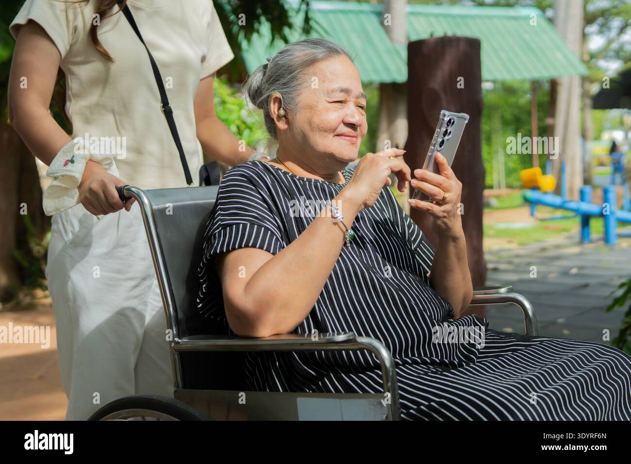 Frau und ältere Mutter im Park, Rollstuhl und Glück vor Freude. Familie, reife Eltern, die das Telefon benutzen, und Tochter, die sich im Freien mit einem smil entspannt Stockfoto