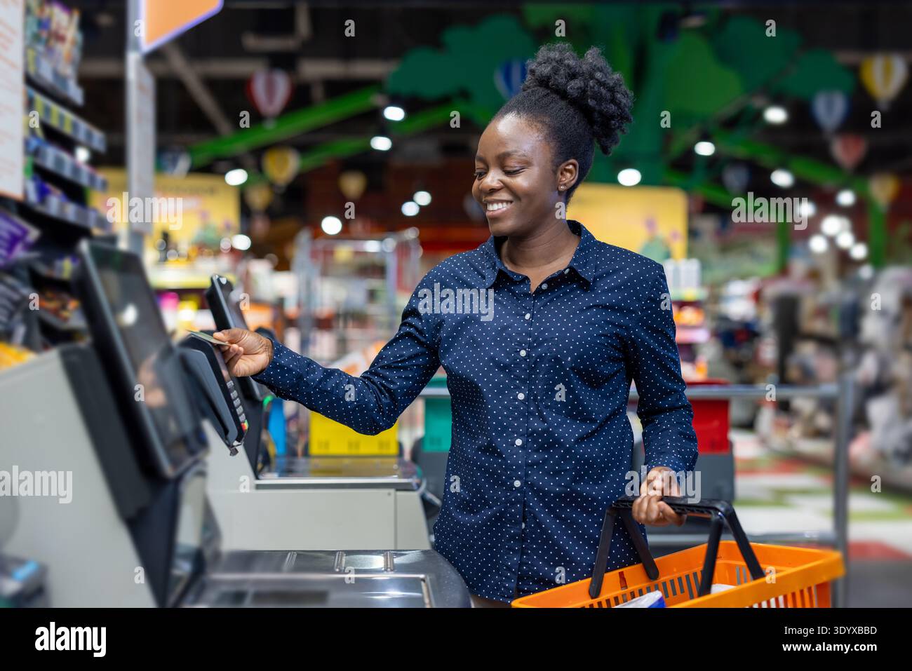Glückliche Frau lächelt, während sie einen Kauf mit einer Kreditkarte an einem automatisierten Selbstkassenterminal in einem geschäftigen Supermarkt abschließt und einen orangefarbenen Warenkorb hält Stockfoto