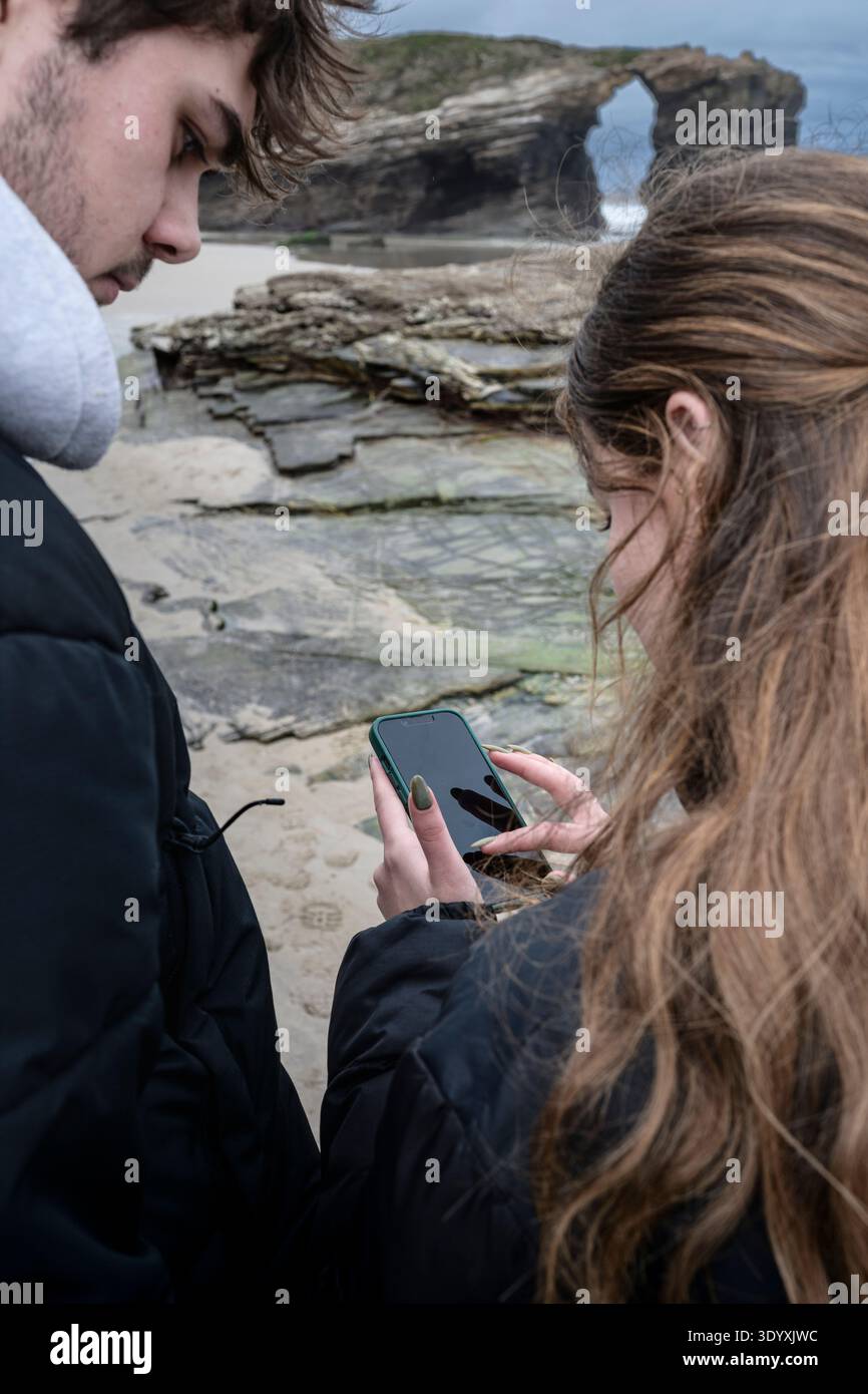 Junge Männer und Frauen am Strand von AS Catedrais blicken auf ein Smartphone, das moderne Reisen, Navigation, digitale Kommunikation und Erfahrungsaustausch anzeigt Stockfoto