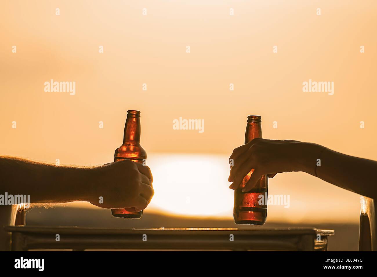 Bier in der Hand eines Paares, der den Sonnenuntergang beobachtet. Zwei Liebende oder Freunde, Mann oder Frau, beobachten den Sonnenuntergang mit Orangenmeer, während sie ein Bier in der Hand halten Stockfoto