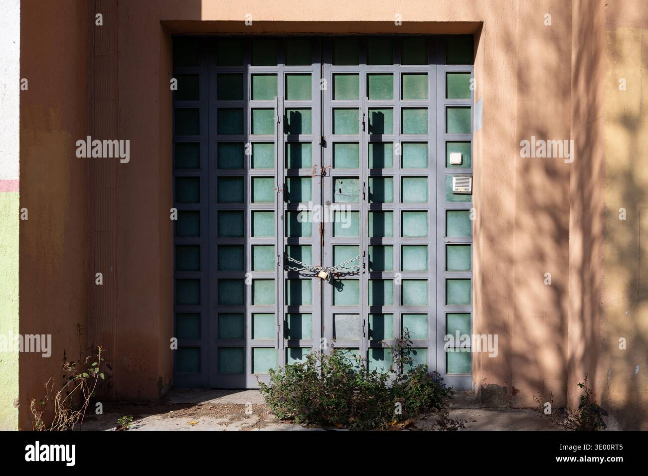 Eine geschlossene Gittertür aus Metall, die mit einem Vorhängeschloss und einer schweren Kette gesichert ist, mit mattierten grünen Glaspaneelen und bewachsenem Unkraut am Eingang eines abando Stockfoto