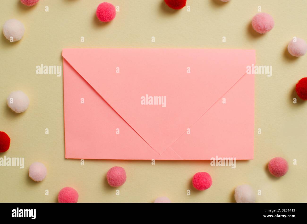Eine pinkfarbene Postkarte auf hellgelbem Hintergrund, umgeben von flauschigen Pompons in Rosa-, Rot- und Weißtönen. Ein festliches und zartes Stillleben Stockfoto