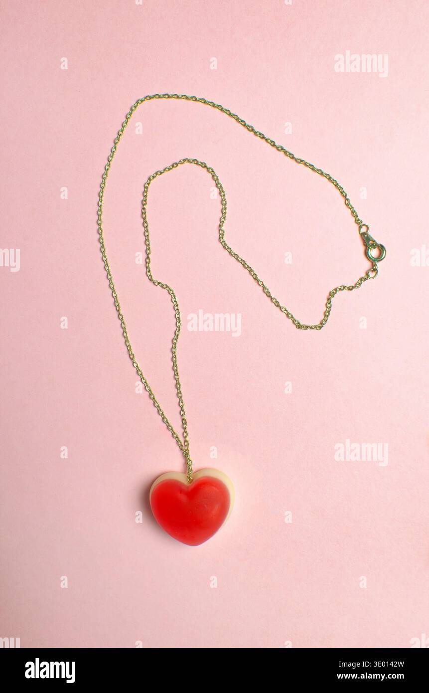 Goldener Kettenschmuck mit rotem Herzgelee auf weichem rosa Hintergrund. Romantische und festliche Komposition zum Thema Liebe und Valentinstag Stockfoto