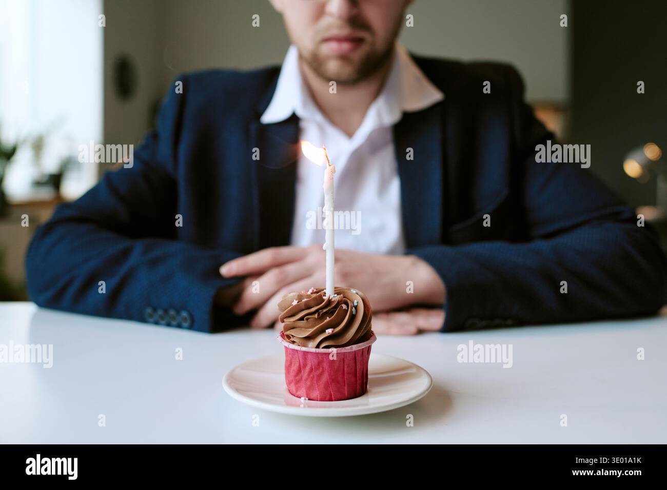 Junger kaukasischer Mann, der allein sitzt und Cupcake mit brennender Kerze anstarrt Stockfoto
