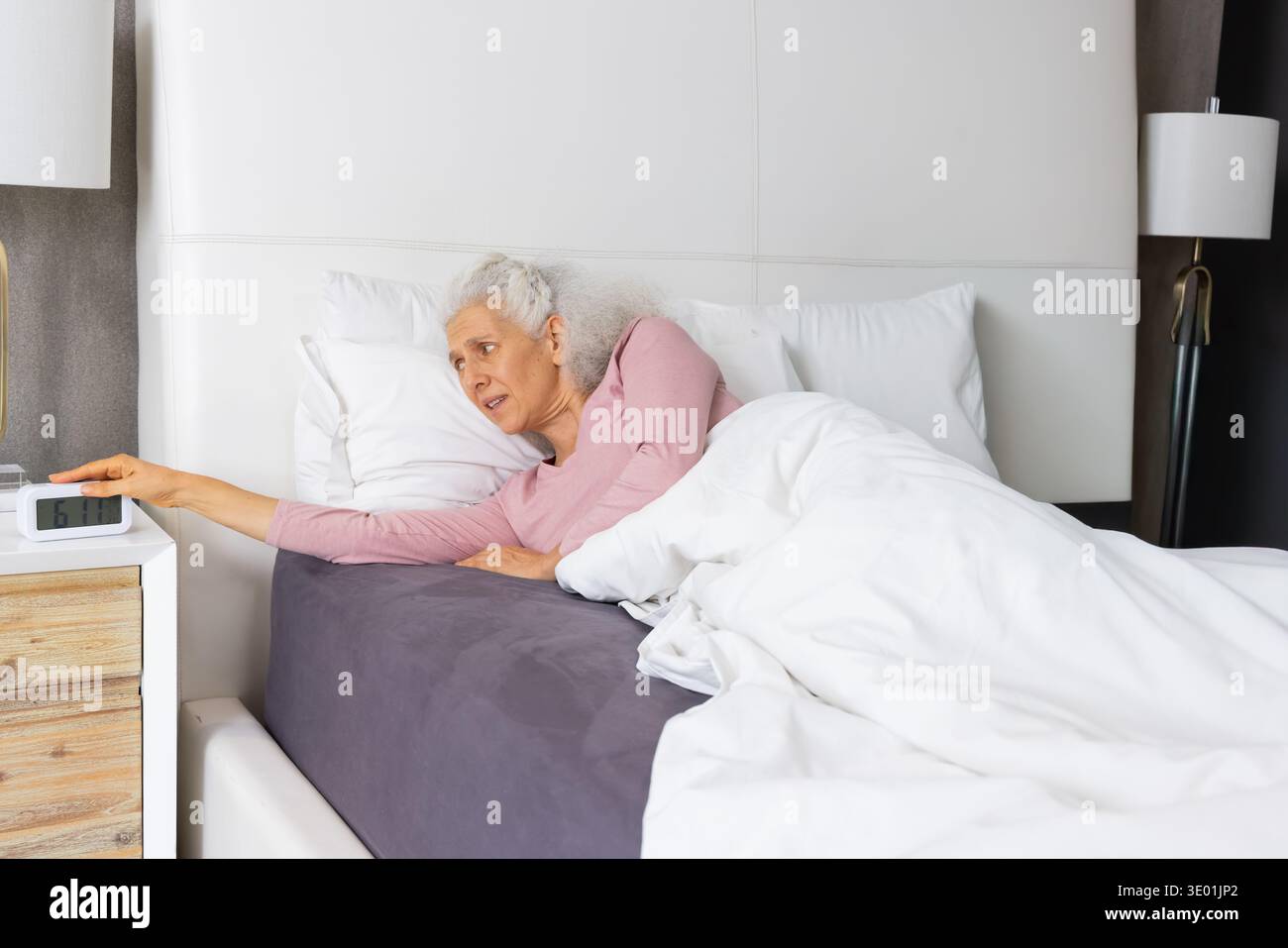Ältere Frau, die im Bett liegt und auf dem linken Ellenbogen nach einem digitalen Wecker auf dem Nachttisch streckt. Schlafzimmer, ältere Menschen, gemütlich, Morgen, sanftes Licht, Federdecke, pi Stockfoto