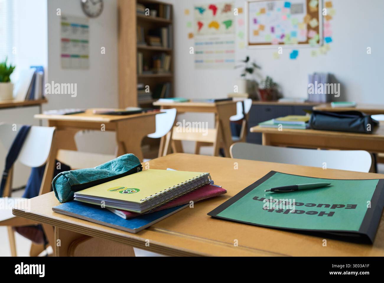 Leeres Klassenzimmer mit Holzschreibtischen mit Notizbüchern, Ordnern mit der Aufschrift Gesundheitserziehung, Stift- und Bleistiftetui auf der Oberfläche angeordnet, Bücherregale und Bildungsplakate sichtbar im Hintergrund Stockfoto