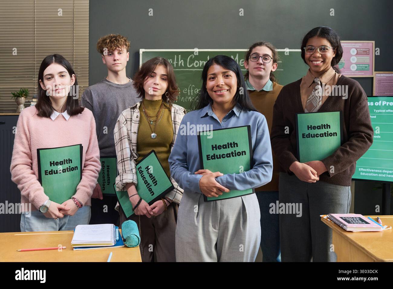Gruppe multiethnischer Teenager, die im Klassenzimmer stehen, Gesundheitsaufklärungsmappen halten, lächelnd und mit der Kamera zugewandt, Tafel mit Gesundheitsthemen im Hintergrund, vielfältige Geschlechterdarstellung Stockfoto