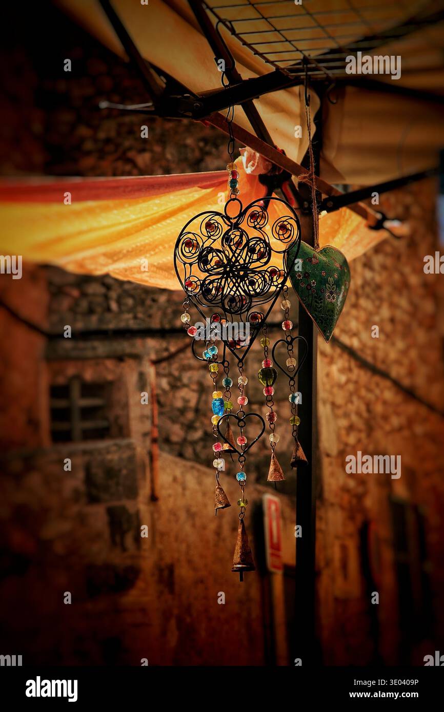 Dekoratives Metall-Herz-Ornament mit bunten Glasperlen, die an einem Marktstand mit warmem Licht hängen und rustikalen Steinhäusern im Hintergrund. Stockfoto