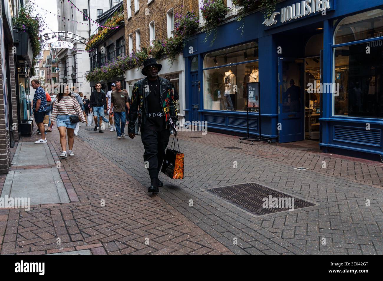 London, Großbritannien - 27. August 2023: Eine lebhafte Straßenszene in Soho, London, mit einem stilvoll gekleideten Mann, der an Geschäften und Fußgängern vorbeiläuft. Stockfoto