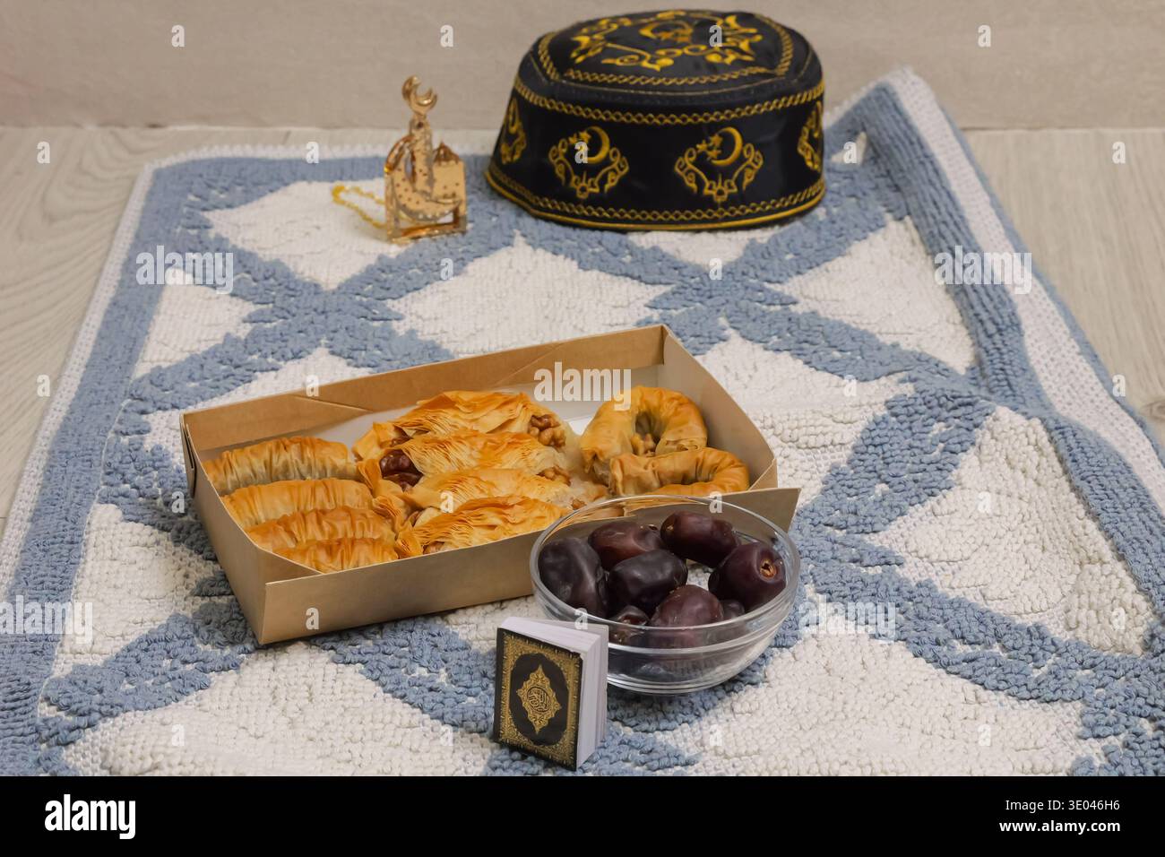 Eine Schachtel Gebäckstücke Auf Einem Gebetsteppich Neben Schwarzgold-Kufi Und Dekorativem Token, Eine Glasschale Mit Datteln Und Weiches Natürliches Licht Schaffen Intime Reflektoren Stockfoto