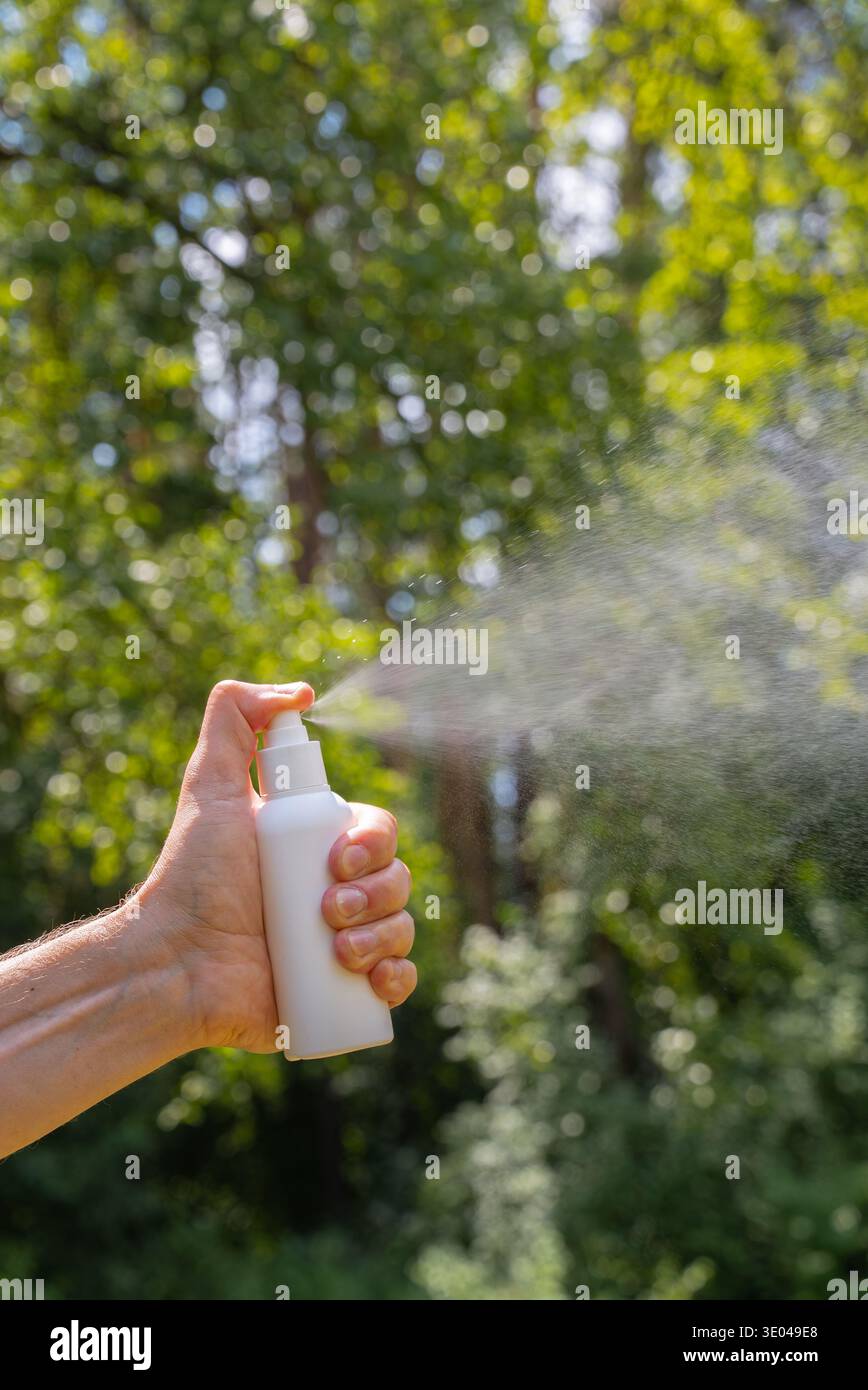 Eine Hand hält eine weiße Flasche und sprüht Insektenschutzmittel vor einem verschwommenen grünen Hintergrund in die Luft. Ideal zum Schutz der Haut vor Insekten während der Fahrt Stockfoto