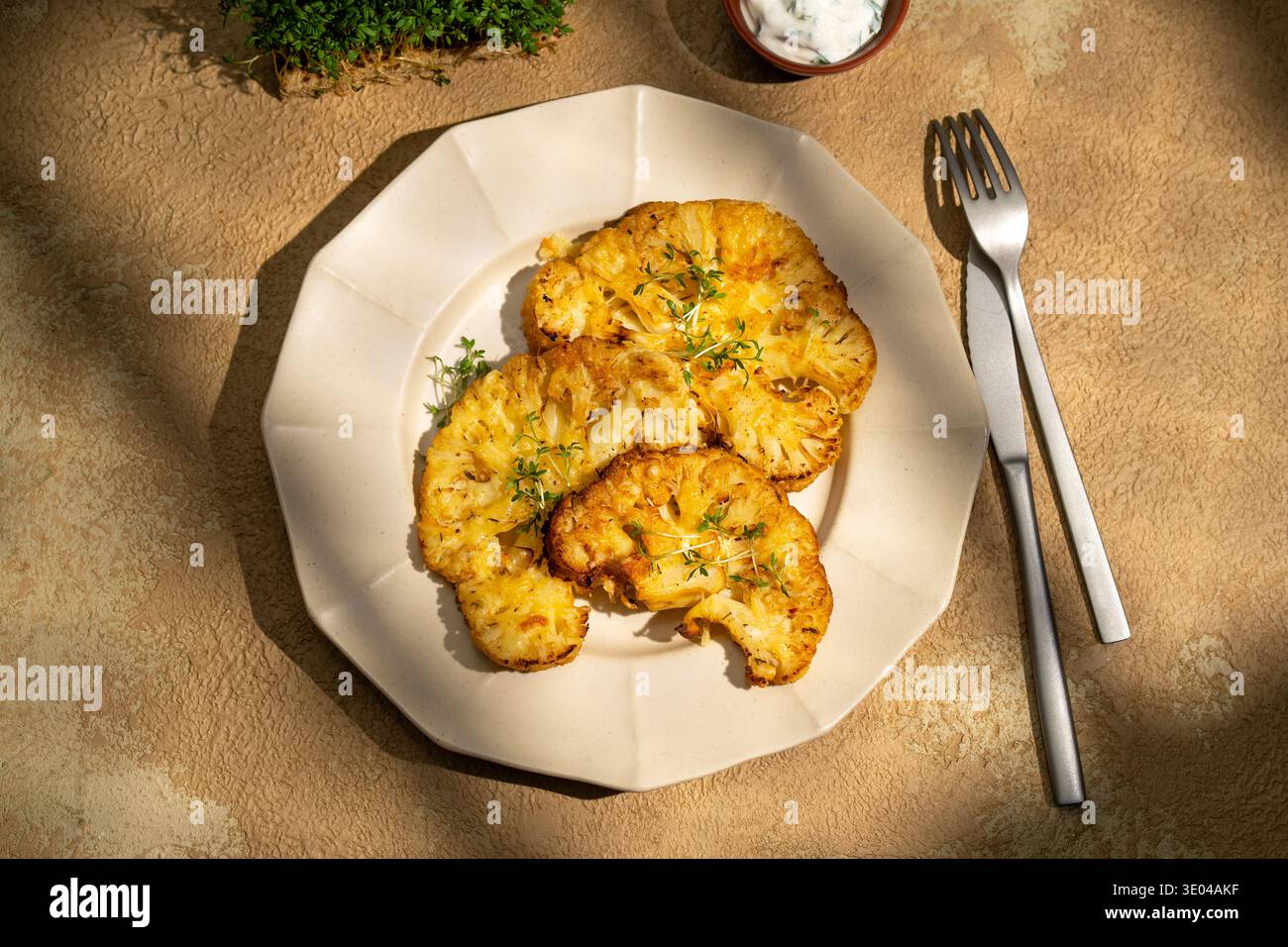 Herzhafte gegrillte Blumenkohlsteaks auf einem Teller serviert, garniert mit frischen Kräutern und begleitet von einer Gabel und einem Messer, die ein gesundes, aromatisches p Stockfoto