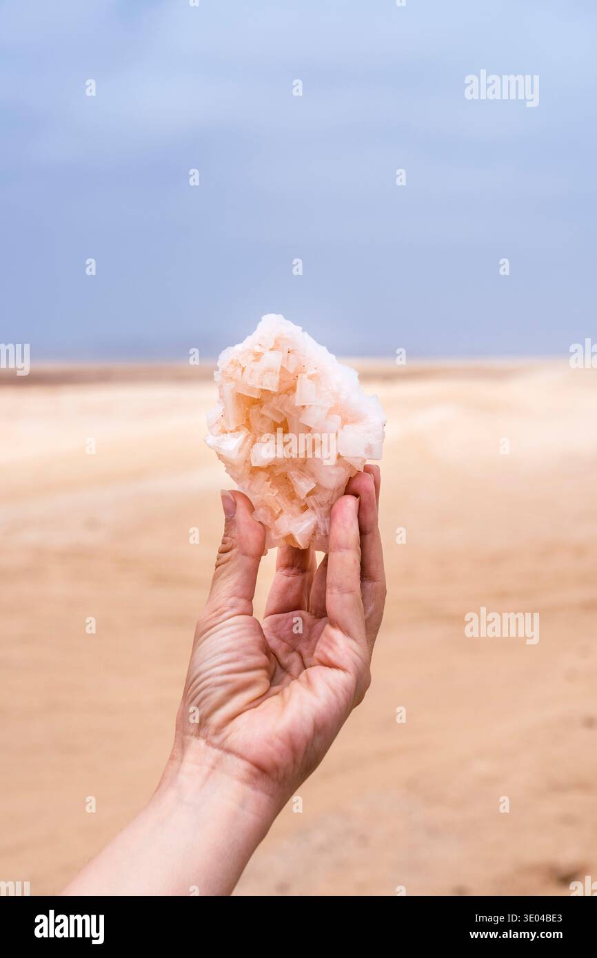 Eine Hand, die einen rohen Salzsteinkristall vor dem Hintergrund der Skeleton Coast in Namibia hält. Der Kristall zeigt komplizierte natürliche Formationen darunter Stockfoto