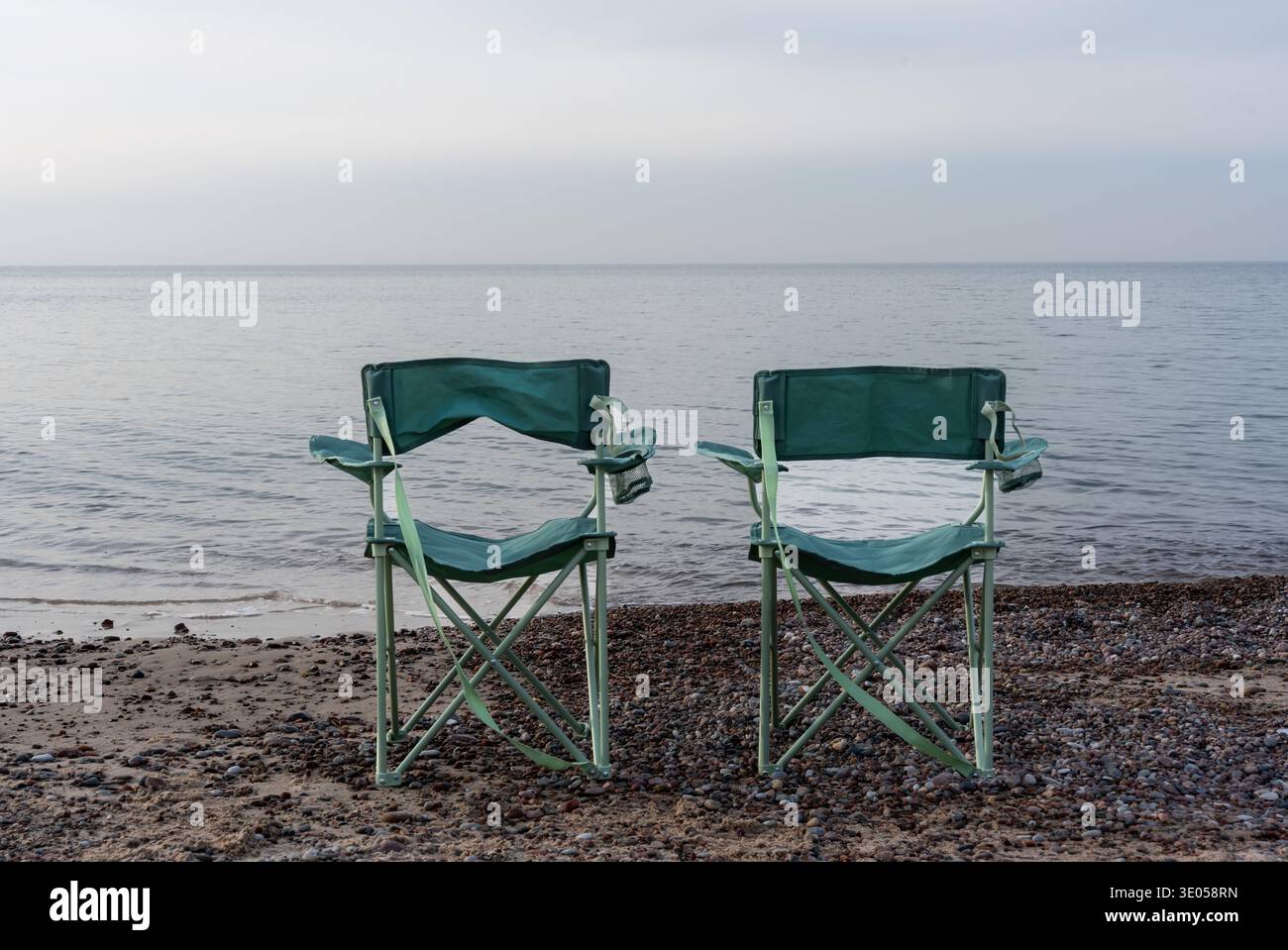 Zwei leere grüne klappbare Campingstühle stehen nebeneinander am Kiesstrand mit Blick auf das ruhige nebelige Meer an ruhigen Tagen. Stockfoto