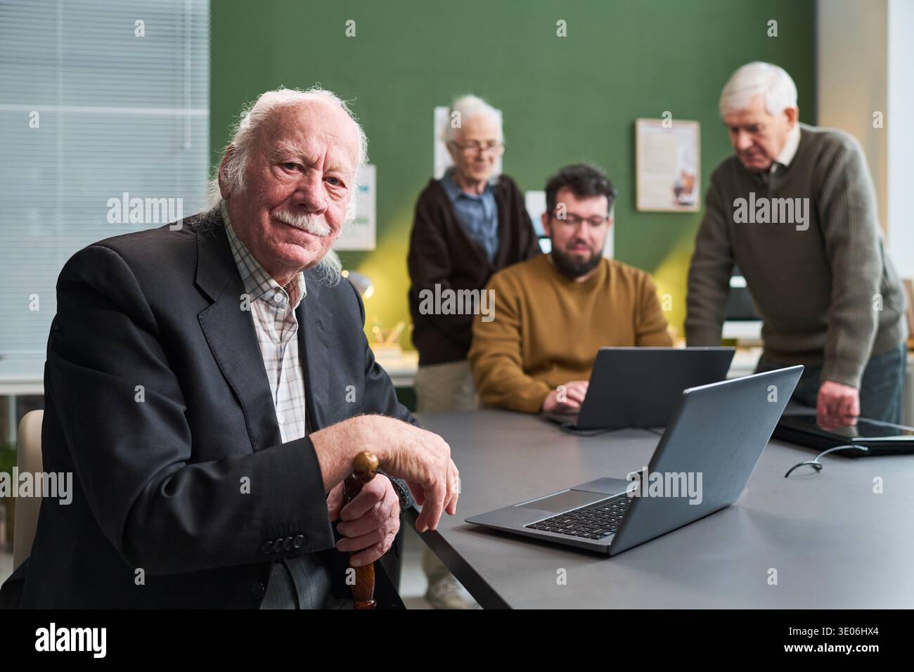 Ein älterer kaukasischer Mann sitzt am Tisch und hält einen Stock, während eine ältere kaukasische Frau, ein älterer kaukasischer Mann und ein junger erwachsener kaukasischer Mann an einem Computerkurs mit Laptops teilnehmen Stockfoto