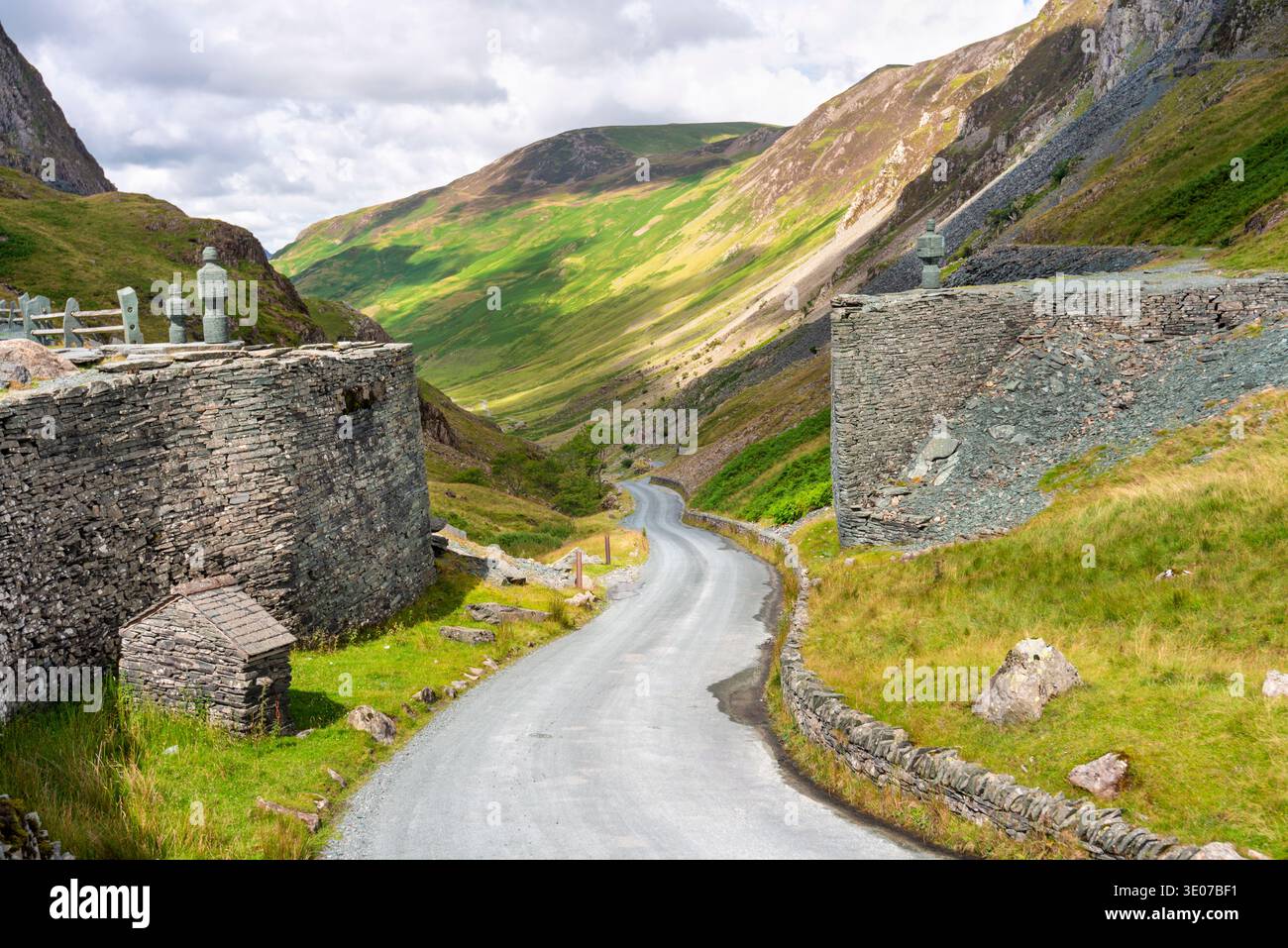 Die gewundene Straße, die zwischen Schiefermauern mit Schieferskulpturen menschlicher Figuren verläuft, schlängelt sich hinunter ins Tal und in die Berge, auf einem warmen, sonnigen Augu Stockfoto