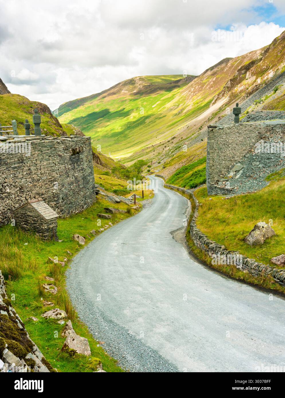 Die gewundene Straße, die zwischen Schiefermauern mit Schieferskulpturen menschlicher Figuren verläuft, schlängelt sich hinunter ins Tal und in die Berge, auf einem warmen, sonnigen Augu Stockfoto