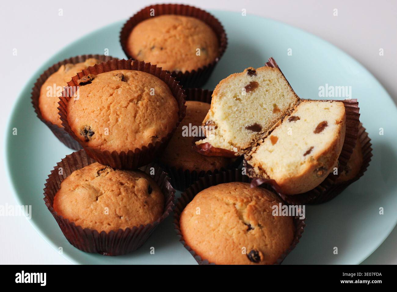 Platte mit hausgemachten Rosinenmuffins, gebacken in braunen Papiereinlagen, wobei ein aufgeschnittener Muffin weiche Schwammbrösel und Rosinen zeigt Stockfoto