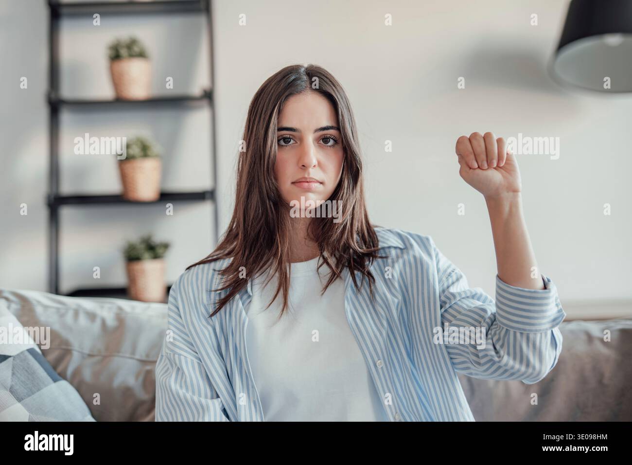 Eine junge Frau sitzt in ihrem Wohnzimmer und zeigt die feministische Geste mit einem traurigen und besorgten Ausdruck. Sie reflektiert Solidarität, Bewusstsein und Einfühlungsvermögen Stockfoto
