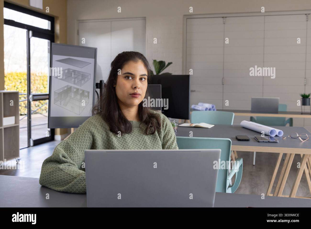 Frau, die am Schreibtisch sitzt und einen Laptop mit großem Monitor benutzt, der 3D-Pläne und -Pläne zeigt Stockfoto