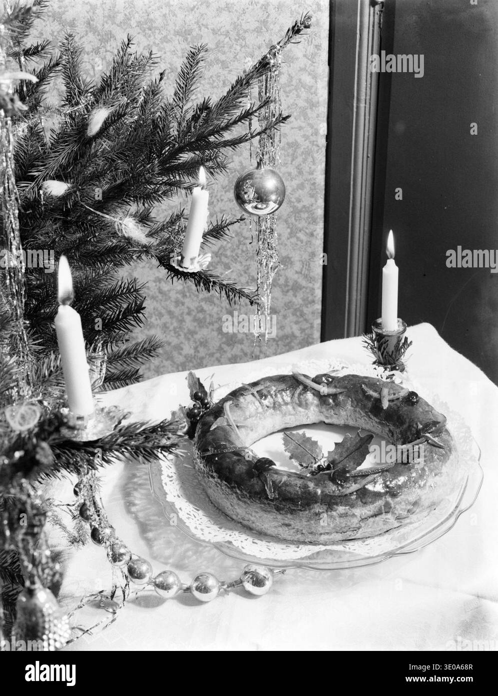 Das Foto zeigt einen traditionellen niederländischen Banketkrans, ein ringförmiges Gebäck aus Blätterteig und Mandelpaste, das während der Weihnachtszeit neben einem geschmückten Weihnachtsbaum ausgestellt wird. Die Banketkrans sind ein festlicher Leckerbissen, der in den Niederlanden häufig während der Weihnachtsfeier serviert wird. Bilder wie diese spiegeln heimische Urlaubstraditionen und saisonale Speisen wider, die typisch für niederländische Haushalte im frühen 20. Jahrhundert waren. Stockfoto