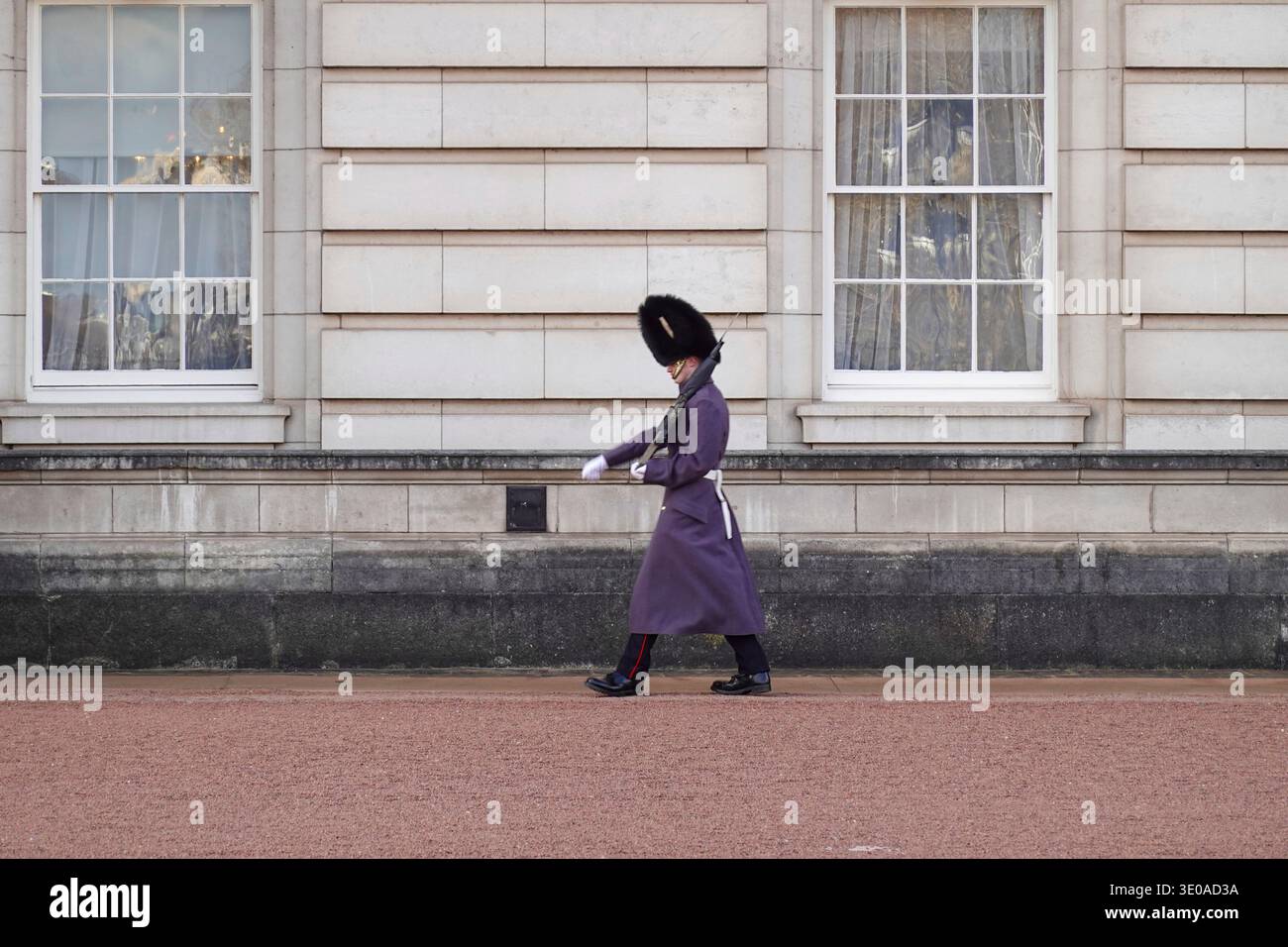 Eine Königsgarde in einem traditionellen grauen Wintermantel und Bärenfellhut marschiert an der Fassade des Buckingham Palace vorbei. London, Vereinigtes Königreich. Stockfoto