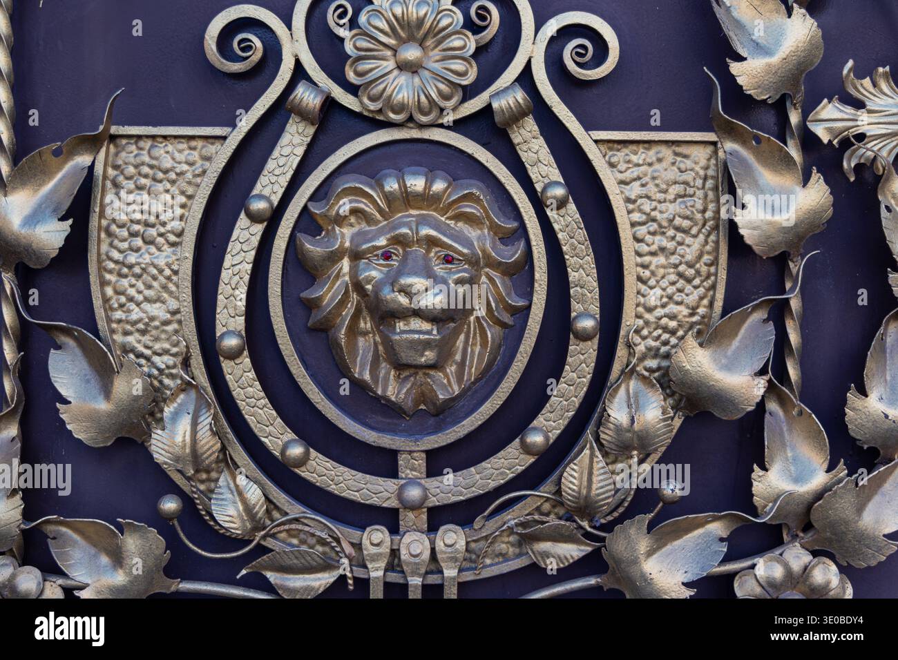 Dekorativer Löwenkopf auf verziertem Metalltor. Nahaufnahme eines geformten Löwenkopfes in einem eleganten Tor aus Schmiedemetall mit floralen Details und strukturierter Struktur Stockfoto