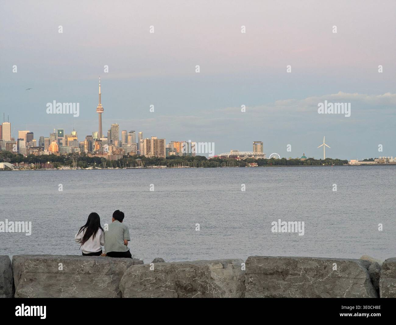 Ein Paar sitzt auf Felsen und blickt auf die Skyline von Toronto mit CN Tower und Windturbine über den Ontario-See bei Sonnenuntergang, Ontario, Kanada. - Smartphone-aufgenommenes Stockfoto