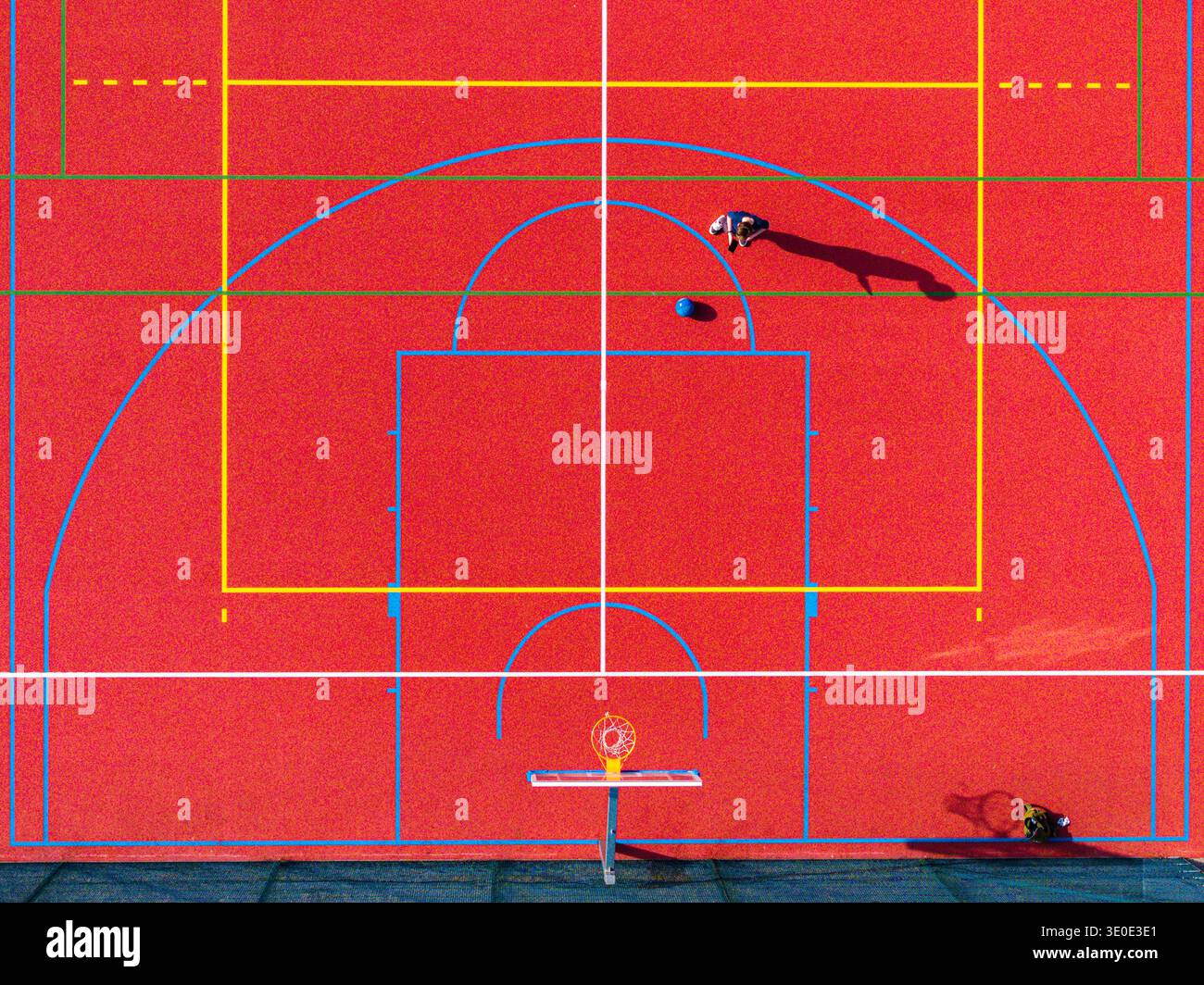 Aus der Vogelperspektive des leuchtend roten Basketballplatzes mit blauen und gelben Linien, einer einsamen Figur und einem Ball, der dynamische Schatten erzeugt, Warschau, Masowische Woiw Stockfoto