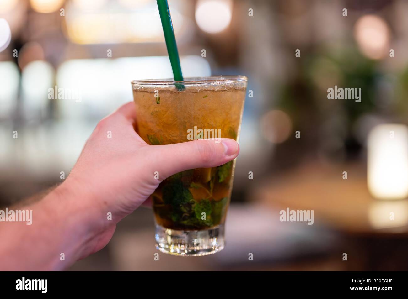 Hand halten ein Glas frischen Mojito-Cocktails mit Eiswürfeln und grünem Stroh. Erfrischendes Sommergetränk mit Rum und Minze in Bar-Atmosphäre, Symbol für Nig Stockfoto
