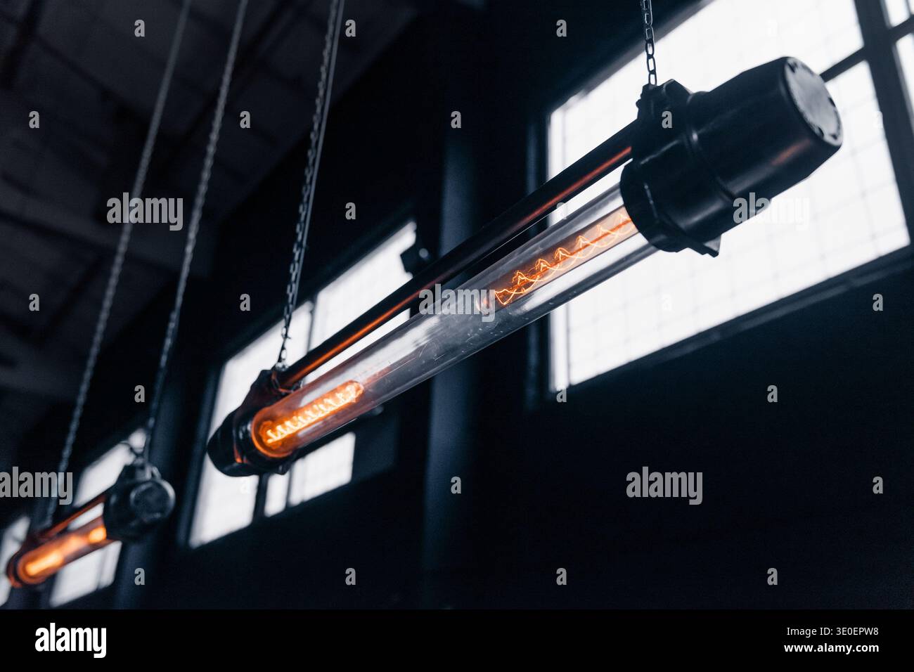 Reihe von industriellen Glühlampen. Eine Niedrigwinkelaufnahme von zwei Hängeleuchten aus röhrenförmigem Filament im Vintage-Stil, die in einer Reihe in einem dunklen Lager hängen. Stockfoto