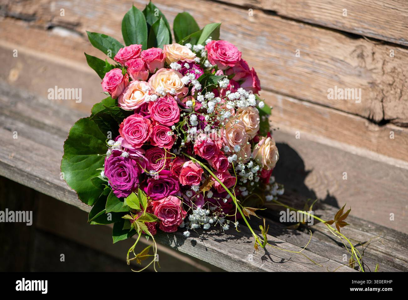 Sonnendurchfluteter Rosenstrauß auf rustikaler Holzbank, hervorgehoben durch warme Schatten und natürliche Texturen in einer Hochzeitsatmosphäre im Freien. Stockfoto