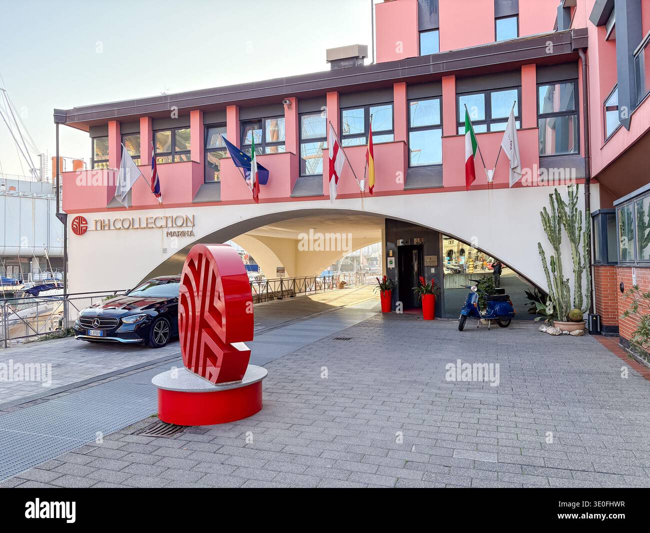 Eingang zum NH Collection Marina Hotel. Genua, Italien. Mit italienischen Flaggen, EU-Flaggen, Skulpturen und geparkten Rollern in der Nähe des Yachthafens. Stockfoto