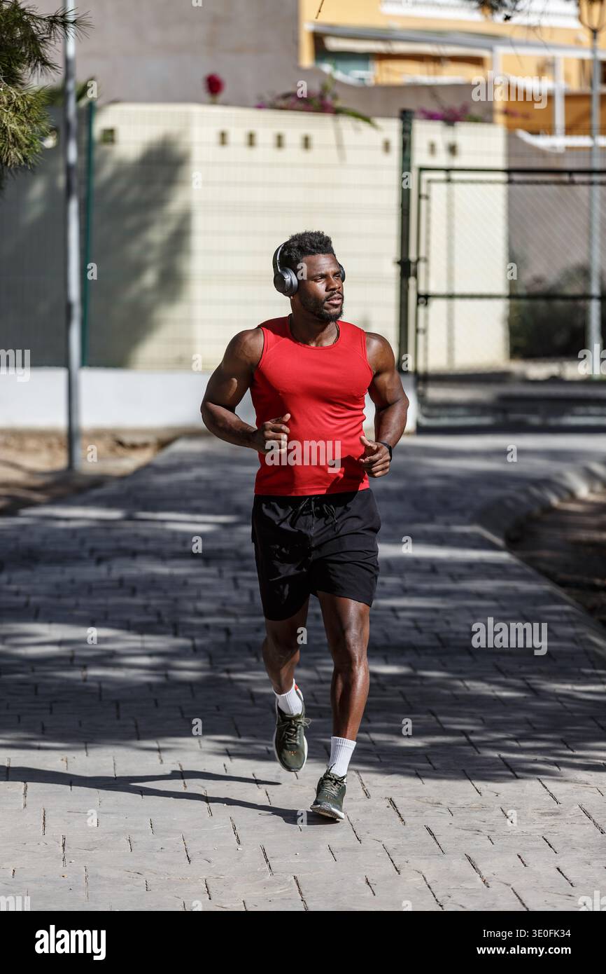 Ein afroamerikanischer Mann joggt durch einen öffentlichen Park, trägt ein rotes ärmelloses Hemd und schwarze Shorts. Er hört Musik mit Kopfhörern, während er es genießt Stockfoto