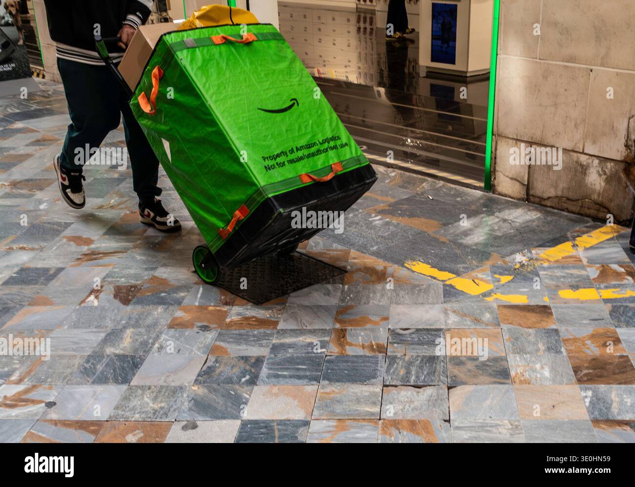 Ein Amazon Logistics-Kurier manövriert einen großen grünen Lieferbeutel auf einem Wagen über eine marmorierte Fußgängerzone in Cartagena, Spanien. Stockfoto