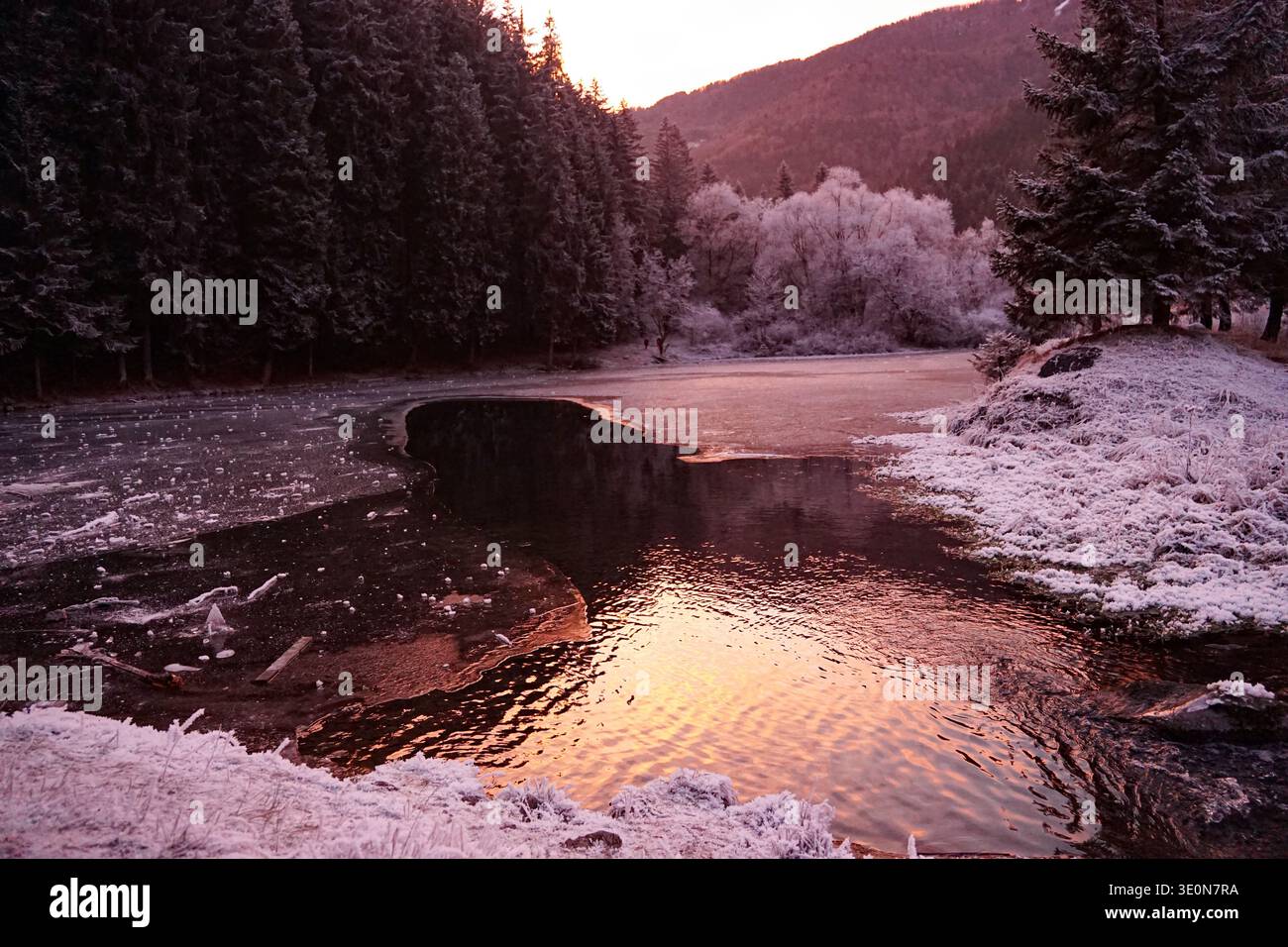 Teilweise gefrorener Bergsee reflektiert das Licht des Sonnenuntergangs mit schneebedeckten Bäumen und bewaldeten Hügeln im Winter Stockfoto