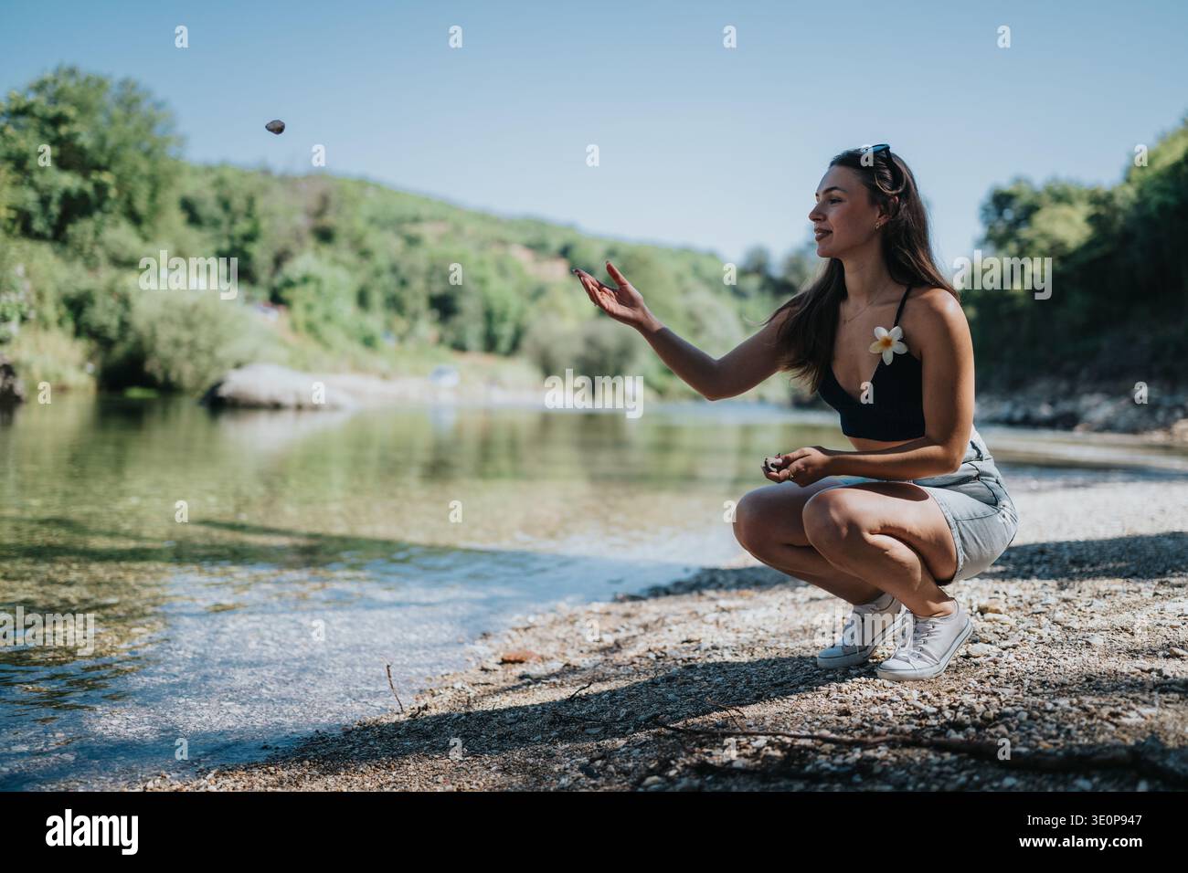Frau, die sich an einem Flussufer hockt und an einem sonnigen Sommertag einen kleinen Stein wirft Stockfoto