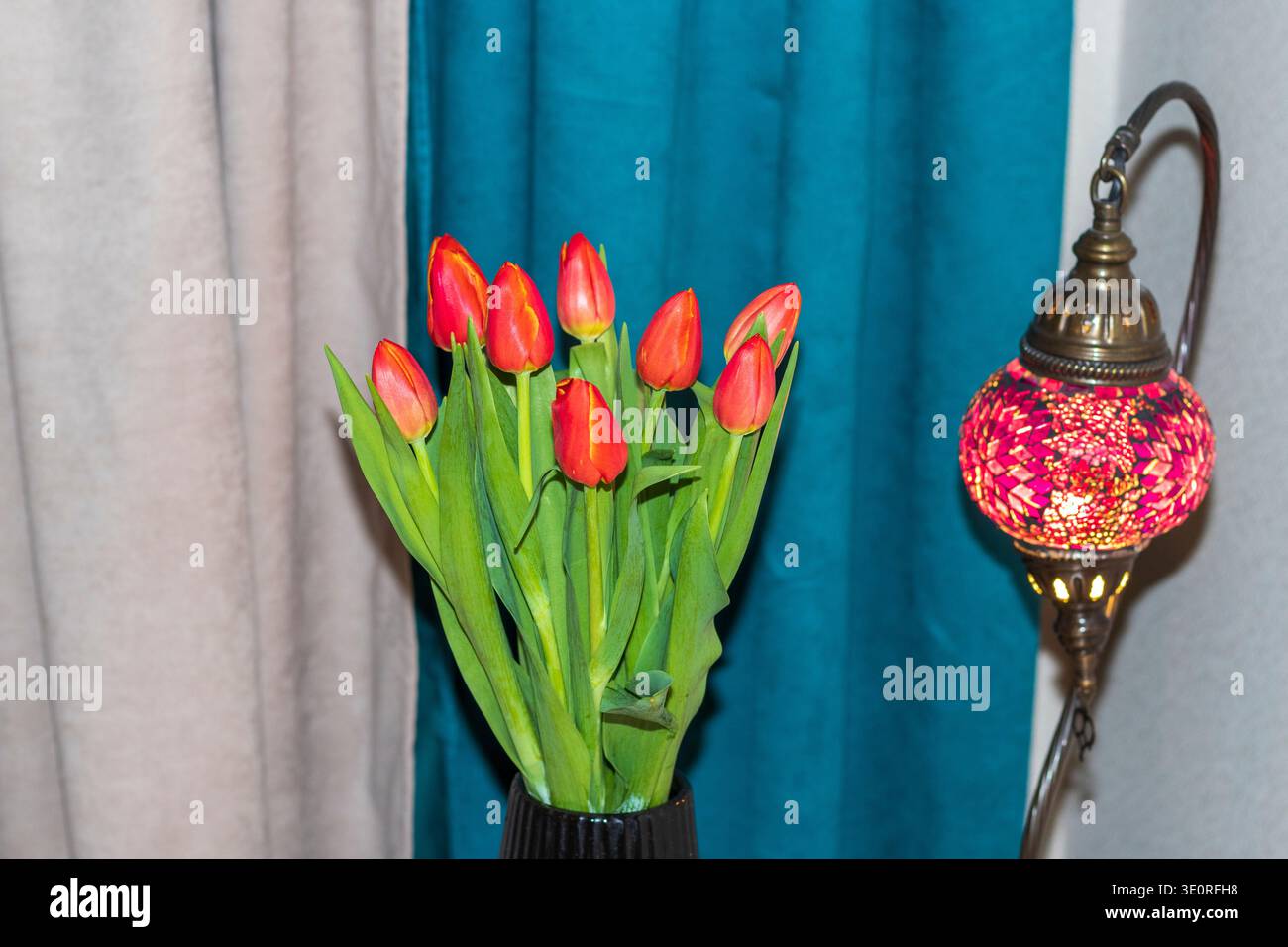 Ein Strauß aus roten Tulpen mit grünen Stielen in einer schwarzen Vase neben einer dekorativen Mosaiklampe im türkischen Stil Stockfoto