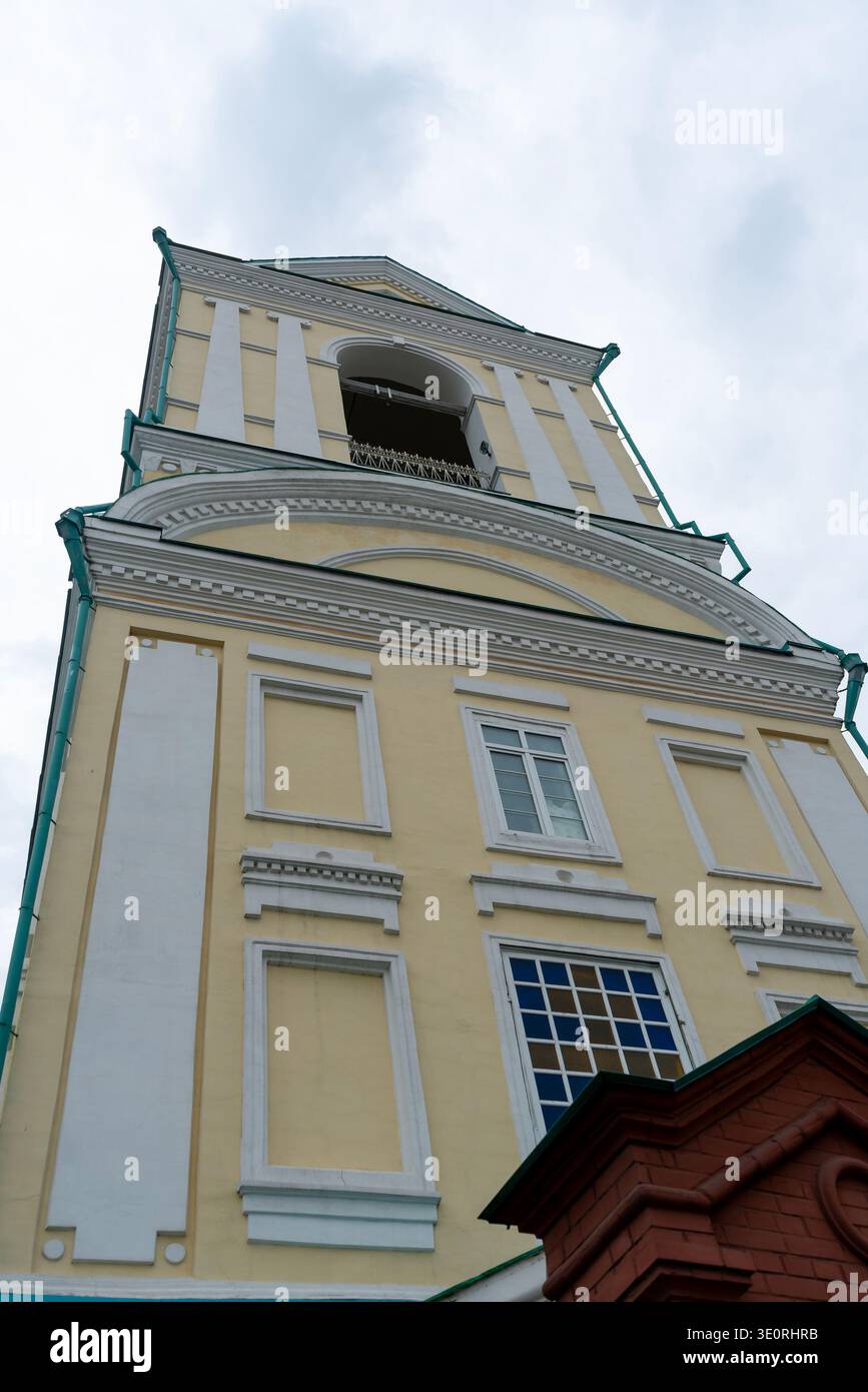 Ein gelb-weißer Kirchturm, der aus einem niedrigen Winkel erfasst wurde und neoklassizistische Details, Säulen und bogenförmige Öffnungen vor einem weichen, bewölkten Himmel zeigt Stockfoto