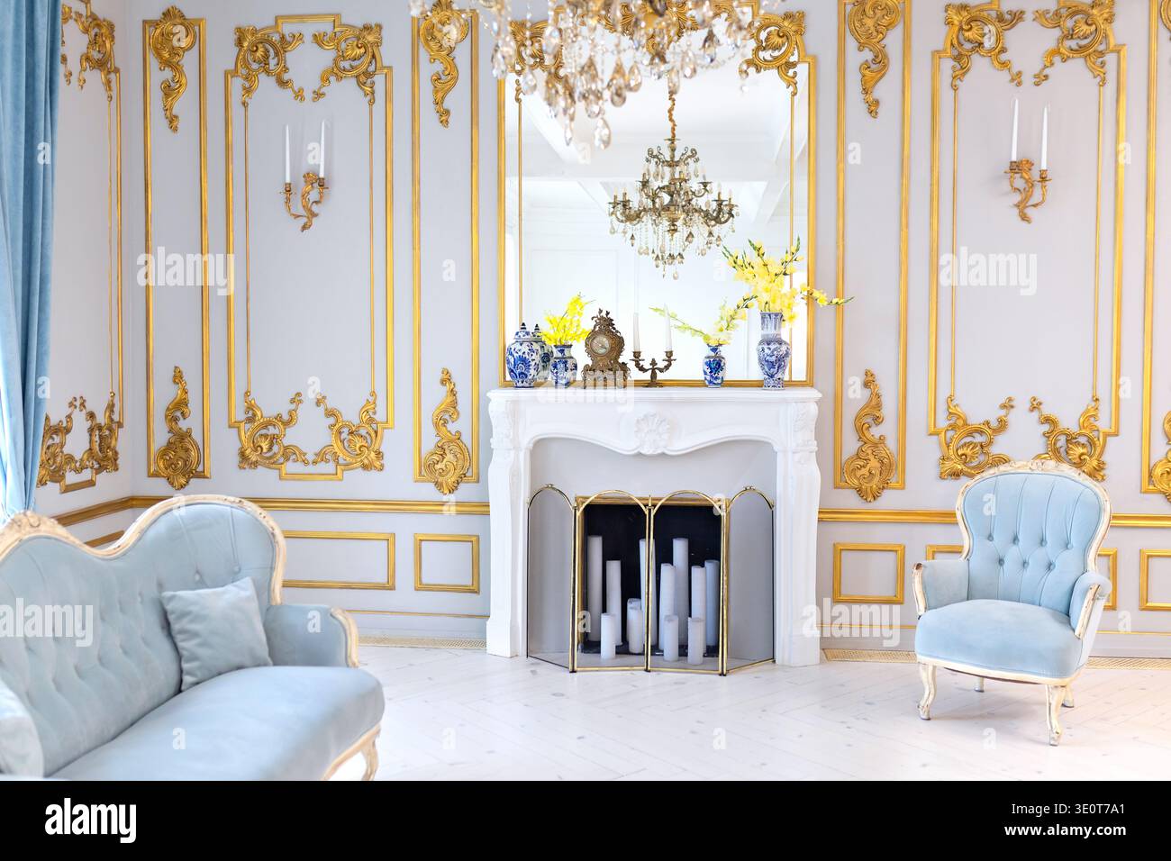 Klassische Einrichtung mit eleganten Sesseln, dekorativem Kamin und Kristallleuchter. Helles Wohnzimmer im Palaststil mit blauen Samtmöbeln Stockfoto