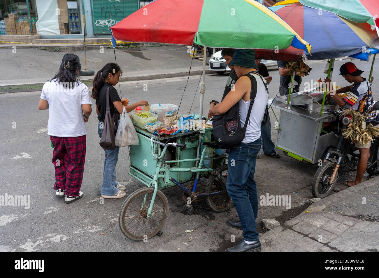 Kunden treffen sich um einen Straßenwagen unter bunten Regenschirmen in Cebu Philippinen, während Händler am späten Nachmittag Snacks zubereiten. Stockfoto